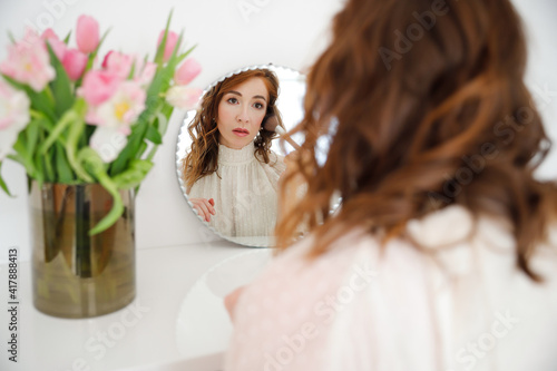 Young pretty woman is doing her makeup in front of the mirror. Red haired woman is preening, primping with a brush and we see her reflection in a mirror