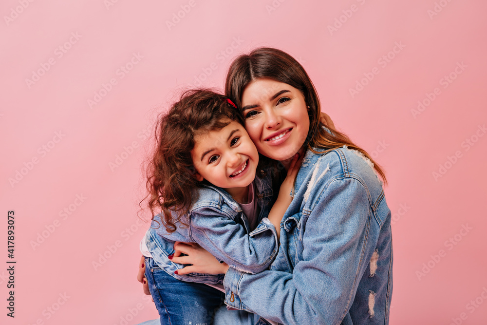 Laughing mother and daughter looking at camera. Front view of young woman with preteen child isolated on pink background. Laughing mother and daughter looking at camera. Front view of young woman with preteen child isolated on pink background.