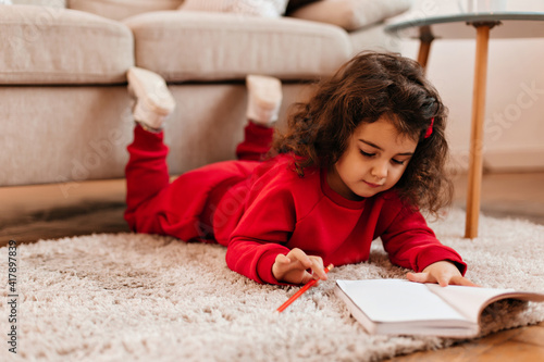 Concentrated kid drawing in notebook. Indoor shot of cute child lying on carpet with pen.