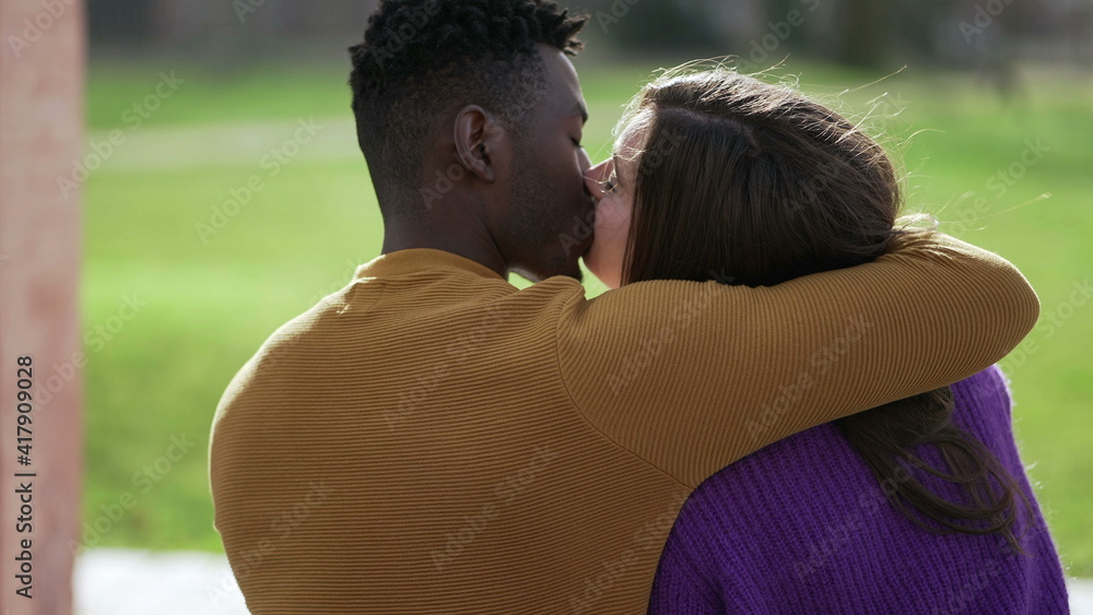 Back of interracial couple kissing sitting at park bench Back of interracial couple kissing sitting at park bench