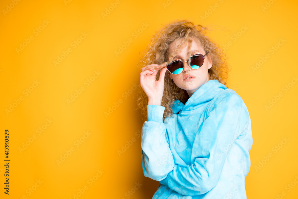 A beautiful girl with blond hair holds glasses with her hand on her face while standing on a bright background A beautiful girl with blond hair holds glasses with her hand on her face while standing on a bright background