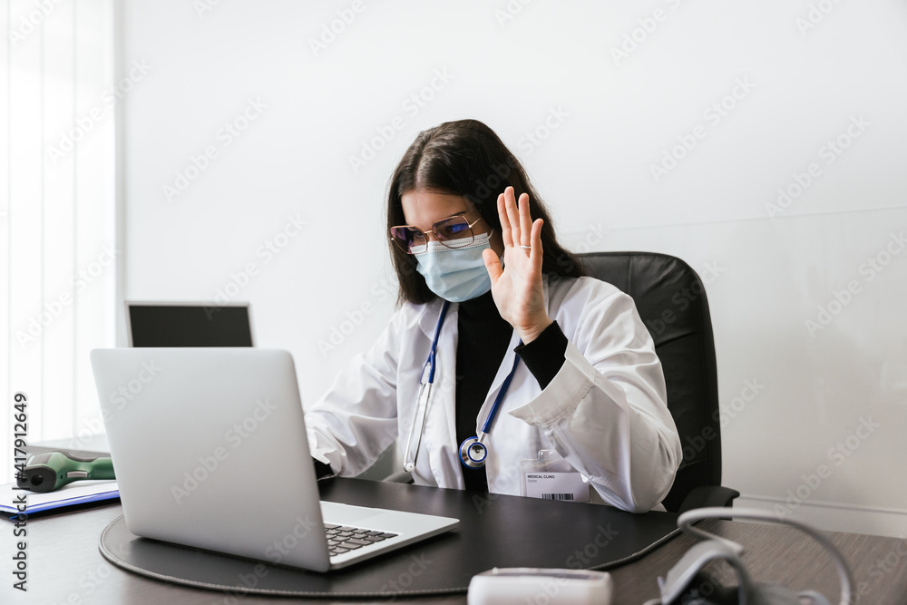 Young doctor working on laptop in office analyzes medical records of his patients infected whit Coronavirus Covid-19 wearing protective face mask - Professional at the desk greets during a video call Young doctor working on laptop in office analyzes medical records of his patients infected whit Coronavirus Covid-19 wearing protective face mask - Professional at the desk greets during a video call