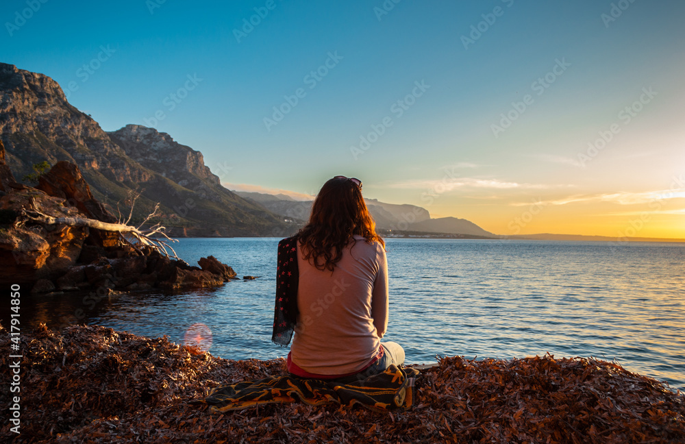 A young and beautiful girl dressed in casual clothes looking at the sunset at a beautiful beach in Artà Mallorca Balearic Islands Spain with a impressive view of the sea and the Tramuntana mountains