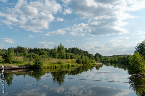 Calm on the river and the reflection of the sky and clouds in the water. Canal and ferry crossing. Ropes are stretched across the river. The banks of the river are overgrown with bushes and trees.