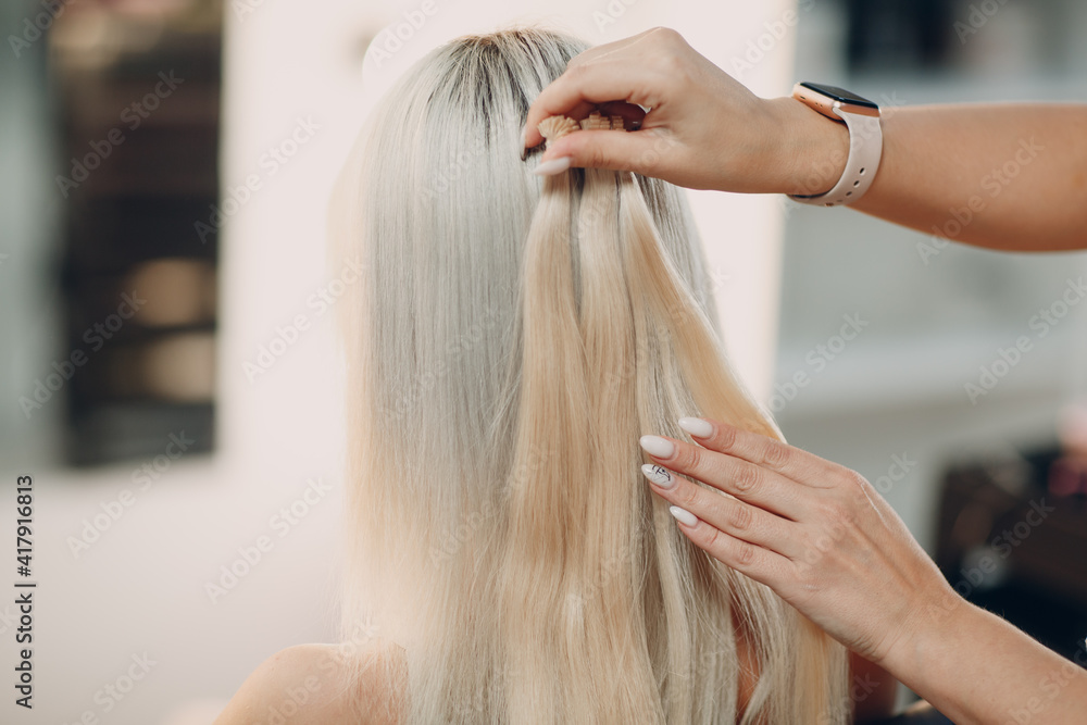 Fototapeta premium Hairdresser female making hair extensions to young woman with blonde hair in beauty salon. Professional hair extension.