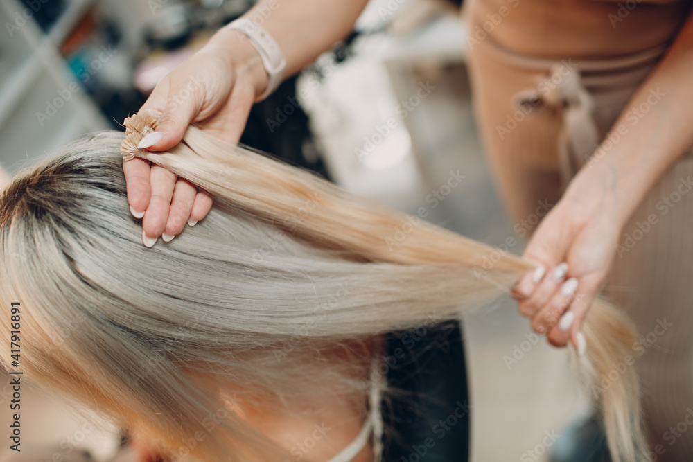 Fototapeta premium Hairdresser female making hair extensions to young woman with blonde hair in beauty salon. Professional hair extension strand of hair.