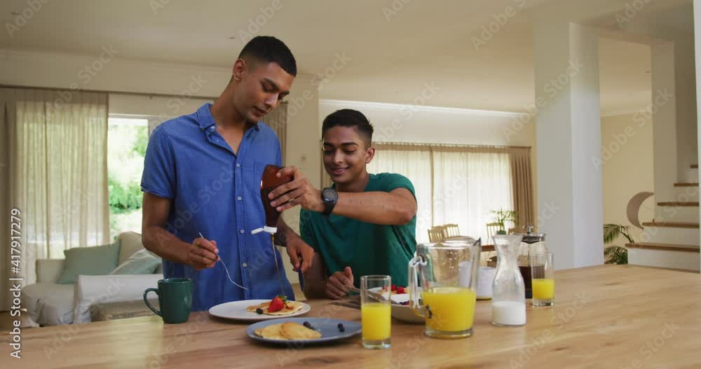 Smiling mixed race gay male couple having pancakes for breakfast in kitchen Smiling mixed race gay male couple having pancakes for breakfast in kitchen