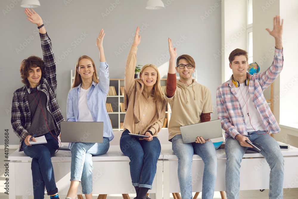 Fototapeta premium Group of happy smiling active students raising hands sitting on desk with digital devices. Young people agree with good idea or answer questions in lecture class. Studying at school or college concept
