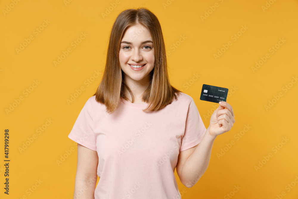 Young caucasian smiling happy rich student woman 20s in casual basic pastel pink t-shirt blank print design holding credit bank card looking camera isolated on yellow color background studio portrait. Young caucasian smiling happy rich student woman 20s in casual basic pastel pink t-shirt blank print design holding credit bank card looking camera isolated on yellow color background studio portrait.