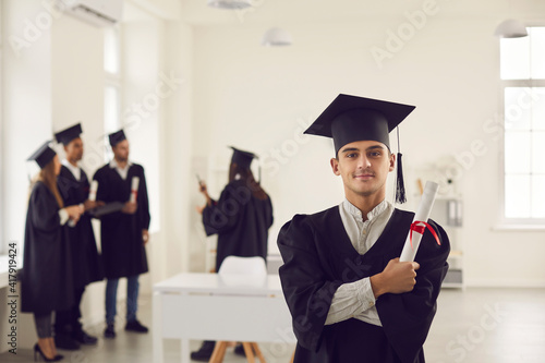 Confident and proud student in the form of a university graduate and with a diploma in hand stands in a bright classroom. Portrait of a guy posing with crossed arms against the backdrop of classmates.