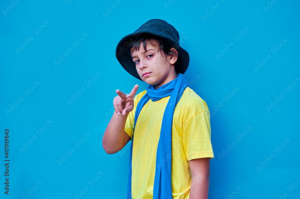 Portrait of defiant boy in yellow t-shirt, scarf and hat doing, on blue background. Copy space Portrait of defiant boy in yellow t-shirt, scarf and hat doing, on blue background. Copy space