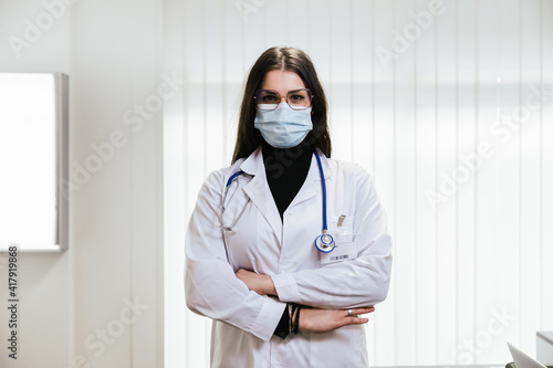 Portrait of young female doctor in medical office at hospital with arms folded wearing protective surgical mask during Coronavirus Covid-19 infection pandemic - Concept of professionalism - Copy space