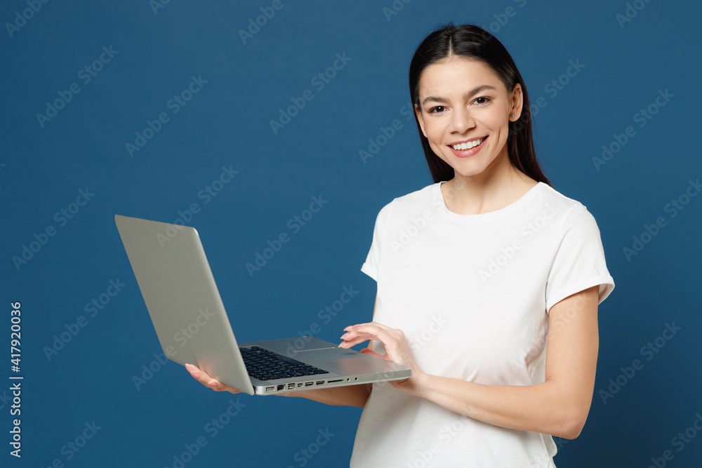 Young smiling cute copywriter freelancer student positive latin woman 20s wear white casual basic t-shirt hold laptop pc computer chatting typing text isolated on dark blue background studio portrait. Young smiling cute copywriter freelancer student positive latin woman 20s wear white casual basic t-shirt hold laptop pc computer chatting typing text isolated on dark blue background studio portrait.