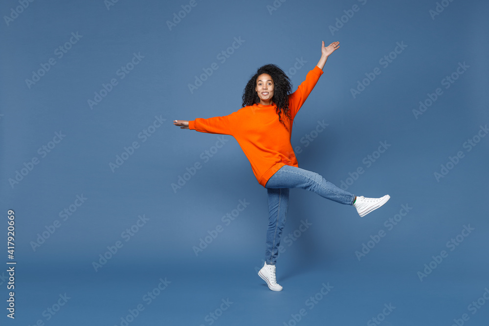 Full length of cheerful smiling young african american woman in casual bright orange sweatshirt dancing rising spreading hands and legs standing on toes isolated on blue background studio portrait.
