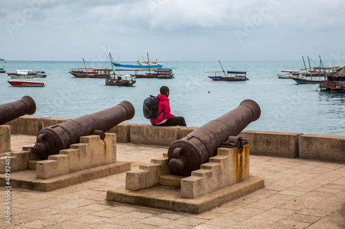 Tanzania, Zanzibar , native tourist boats anchored in a harbor near stone town. Canons are in an 1800's defensive position, boy sitting on sea wall