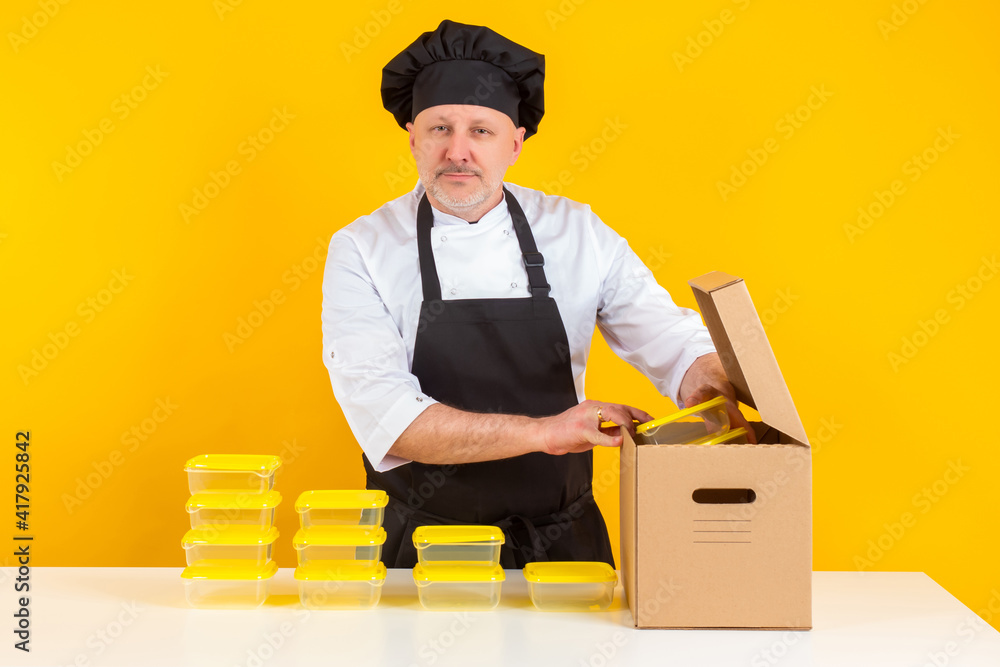 The chef takes plastic containers out of the box. Cooking takeaway food. Serving containers for lunches. The cook looks at the camera on an orange background. The chef takes plastic containers out of the box. Cooking takeaway food. Serving containers for lunches. The cook looks at the camera on an orange background.