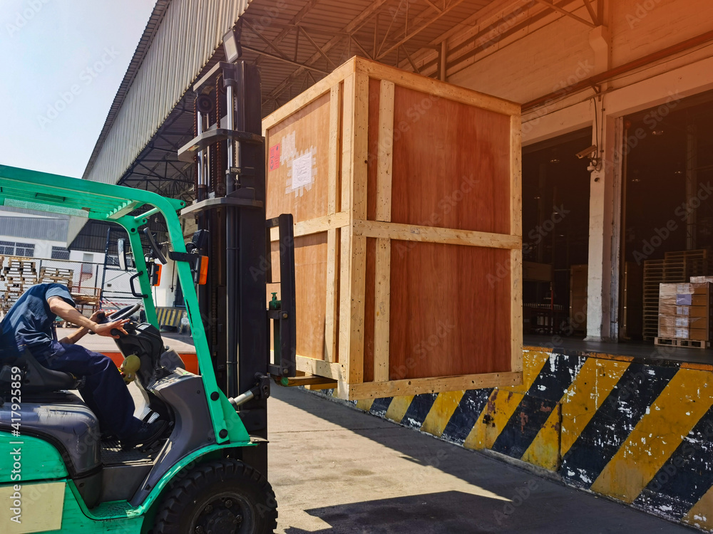 Worker driving forklift loading shipment carton boxes goods on wooden pallet at loading dock from container truck to warehouse cargo storage in freight logistics, transportation industrial, delivery