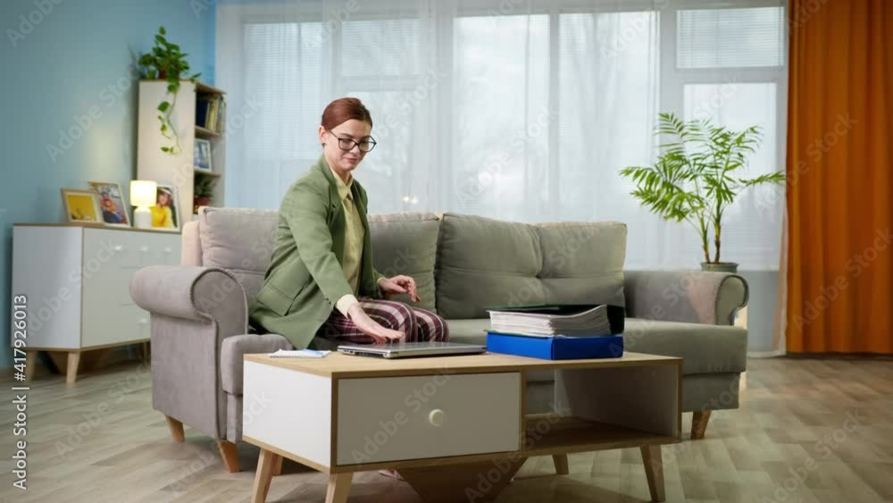 handsome female office worker in pajamas and jacket works remotely at home sitting on the couch with a laptop and documents in his hands, freelance handsome female office worker in pajamas and jacket works remotely at home sitting on the couch with a laptop and documents in his hands, freelance