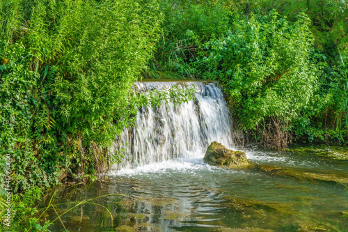 Waterfall in forest. River banks overgrown by solid wall of green bushes and trees
