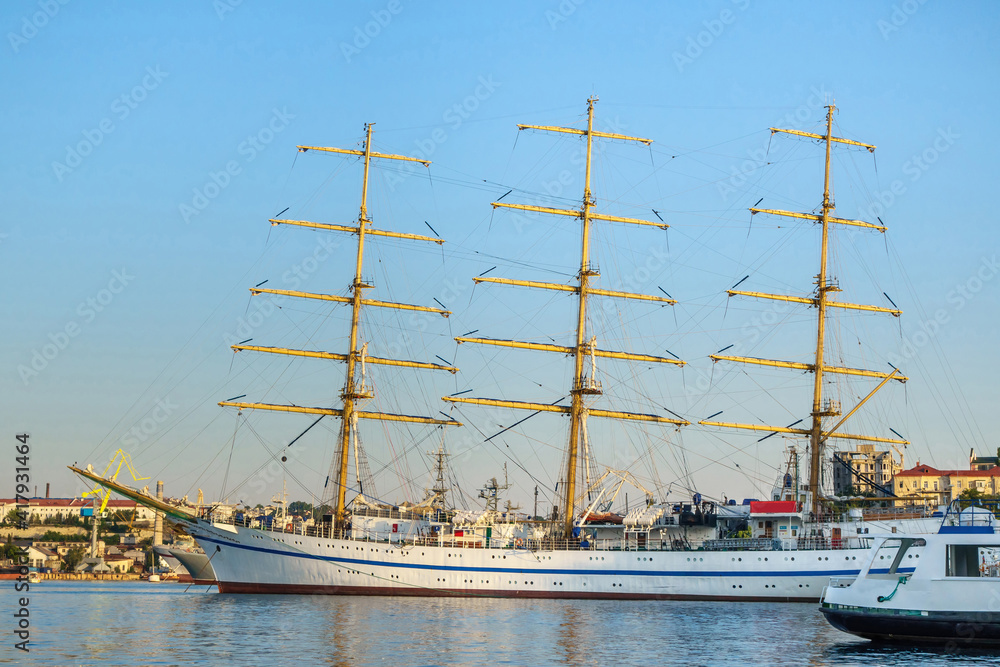 Silhouette of sailing ship in sea harbor among modern boats and barges. Coastal city scape is on background