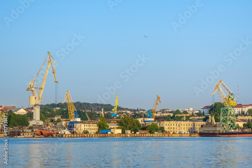 Panorama of industrial part of sea port. There are working cranes, ships being repaired and office buildings. Small figure of seagull on clear sky background