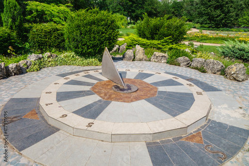 Ancient sundial showing exact time and sides of the world. Dial is surrounded by ornament of stones and blooming plants. Shot in urban public park Salgirka, Simferopol, Crimea. Park founded in 1795