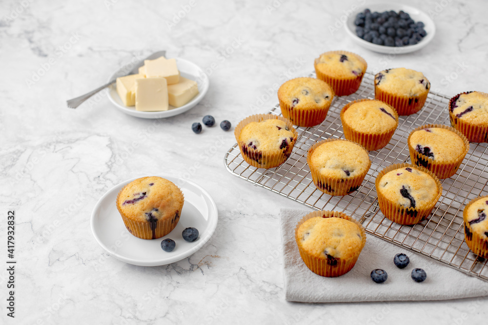 Obraz premium Homemade Blueberry Muffins on a Cooling Rack; One on a White Plate; One on a Gray Napking; Butter on Plate; Blueberries on Plate; Gray and White Marble Countertop