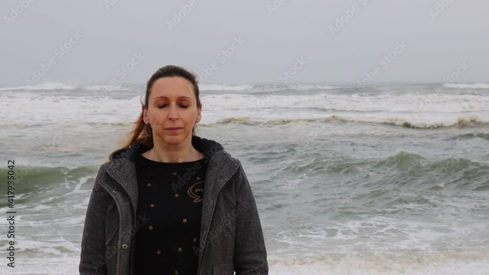 Caucasian woman alone at the beach in winter clothes looking at camera peacefully and closing eyes with ocean waves in the background. Winter ocean landscape. Meditation, relaxation or health concepts Caucasian woman alone at the beach in winter clothes looking at camera peacefully and closing eyes with ocean waves in the background. Winter ocean landscape. Meditation, relaxation or health concepts