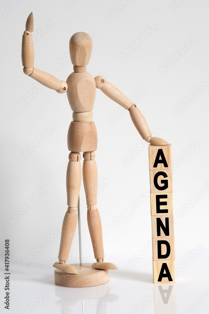 The wooden man raised his hand, and under his left hand he has cubes with the inscription - AGENDA The wooden man raised his hand, and under his left hand he has cubes with the inscription - AGENDA