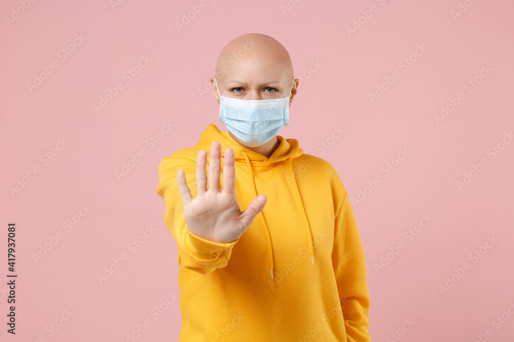 Young bald serious strict woman 20s without hair in yellow casual shirt in sterile face mask to safe coronavirus virus covid-19, quarantine do stop palm gesture refusing isolated on pink background. Young bald serious strict woman 20s without hair in yellow casual shirt in sterile face mask to safe coronavirus virus covid-19, quarantine do stop palm gesture refusing isolated on pink background.