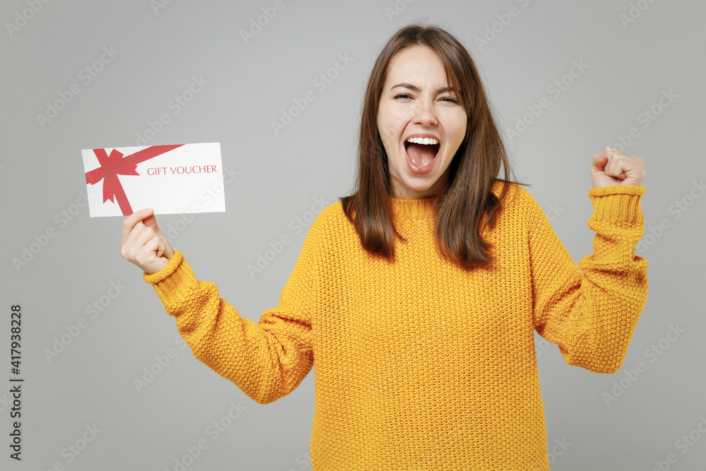 Young excited overjoyed caucasian woman 20s in casual knitted yellow sweater point index finger on gift voucher flyer mock up do winner gesture clench fist isolated on grey background studio portrait Young excited overjoyed caucasian woman 20s in casual knitted yellow sweater point index finger on gift voucher flyer mock up do winner gesture clench fist isolated on grey background studio portrait