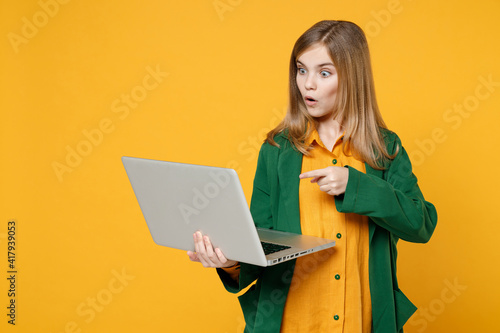 Little blonde pretty cute kid girl 12-13 years old wearing casual clothes point index figer on laptop pc computer isolated on yellow background children studio portrait. Childhood lifestyle concept © ViDi Studio
