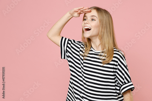 Little blonde kid girl 12-13 years old in striped oversized t-shirt holding hand at forehead looking far away distance isolated on pastel pink background children studio . Childhood lifestyle concept.