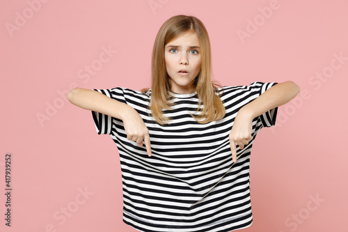 Little blonde smiling kid girl 12-13 years old wearing striped oversized t-shirt point index finger down on copy space area isolated on pastel pink background children. Childhood lifestyle concept. © ViDi Studio