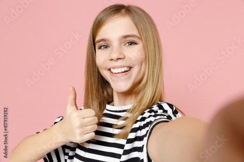 Close up little blonde smiling kid girl 12-13 years old in striped oversized t-shirt do selfie shot on mobile phone show thumb up like gesture isolated on pink background children Childhood concept.