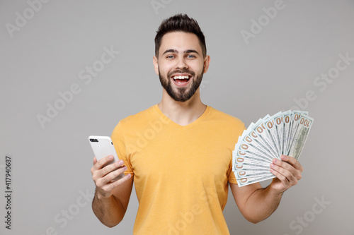 Fotografie Young bearded rich happy man 20s wearing casual yellow basic t-shirt hold mobile cell phone holding fan of cash money in dollar banknotes shopping online isolated on grey background studio portrait