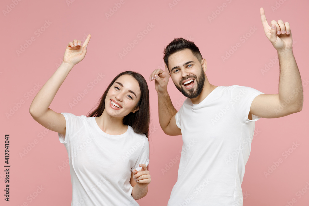 Young cheerful happy couple two friends man woman in white basic blank print design t-shirts posing dancing fooling around celebrate have fun isolated on pastel pink color background studio portrait. Young cheerful happy couple two friends man woman in white basic blank print design t-shirts posing dancing fooling around celebrate have fun isolated on pastel pink color background studio portrait.