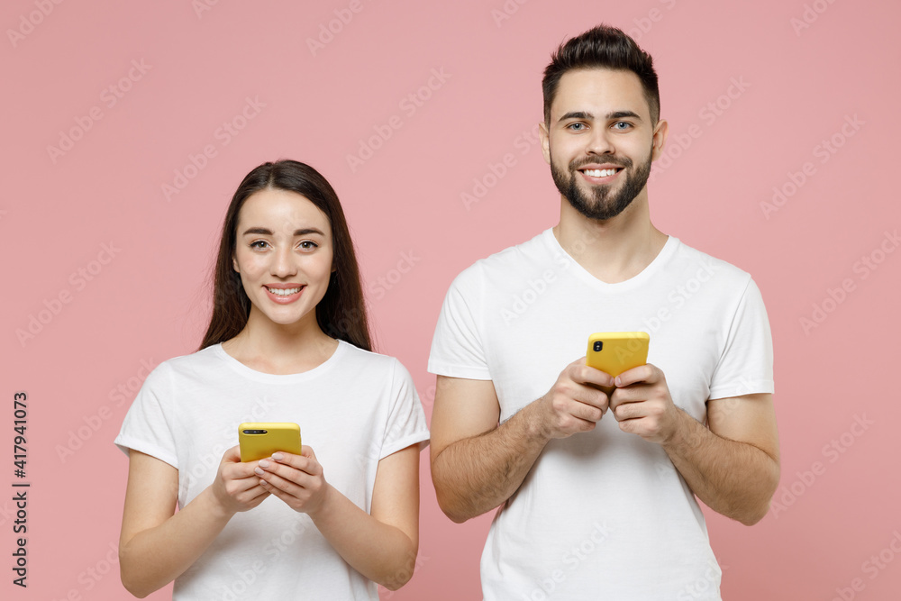 Young couple two friends man brunette woman 20s in white basic blank print design t-shirts hold in hand using mobile cell phone chatting typing sms isolated on pastel pink background studio portrait. Young couple two friends man brunette woman 20s in white basic blank print design t-shirts hold in hand using mobile cell phone chatting typing sms isolated on pastel pink background studio portrait.
