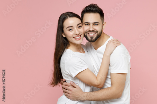 Young happy couple in love two friends bearded man brunette woman 20s in white basic t-shirts looking camera standing smiling hugging embrace isolated on pastel pink color background studio portrait.