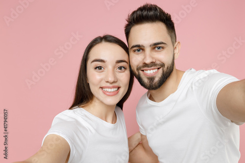Close up young cheerful couple two friends man brunette woman in white basic blank print design t-shirt doing selfie shot on mobile cell phone isolated on pastel pink color background studio portrait