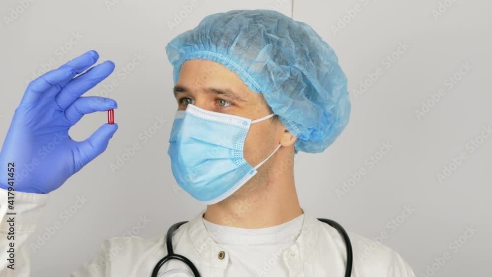 A young handsome doctor in a medical mask holds a red pill in his hands presenting a new drug.A young doctor shows a new drug at the presentation, a red pill for all diseases.