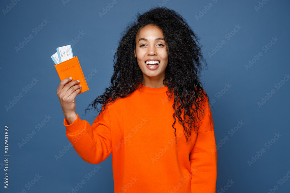 Cheerful young traveler tourist african american woman in orange sweatshirt hold passport tickets isolated on blue background. Passenger travel abroad on weekends getaway. Air flight journey concept.