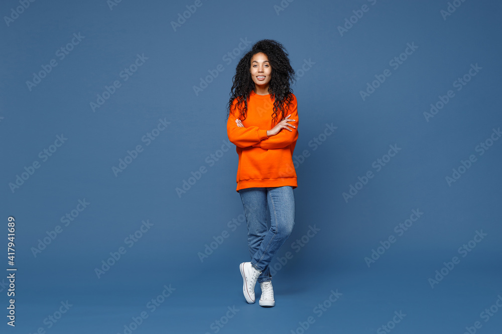 Full length of beautiful smiling young african american woman in casual basic bright orange sweatshirt standing holding hands crossed looking camera isolated on blue color background studio portrait. Full length of beautiful smiling young african american woman in casual basic bright orange sweatshirt standing holding hands crossed looking camera isolated on blue color background studio portrait.