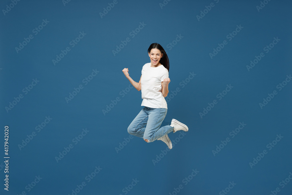 Full length young happy friendly overjoyed brunette latin woman 20s wear white casual basic t-shirt do winner gesture clench fist jumping high isolated on dark blue color background studio portrait Full length young happy friendly overjoyed brunette latin woman 20s wear white casual basic t-shirt do winner gesture clench fist jumping high isolated on dark blue color background studio portrait