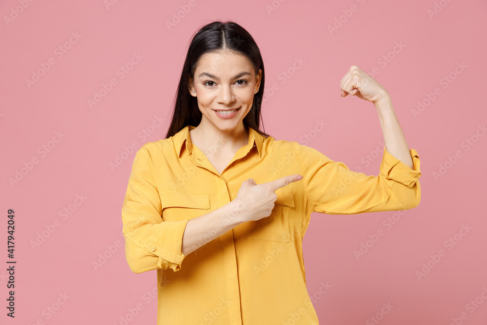 Young brunette cute nice latin strong sporty fitness cute happy woman 20s wearing yellow casual shirt point index finger on biceps muscles on hand isolated on pastel pink background studio portrait. Young brunette cute nice latin strong sporty fitness cute happy woman 20s wearing yellow casual shirt point index finger on biceps muscles on hand isolated on pastel pink background studio portrait.