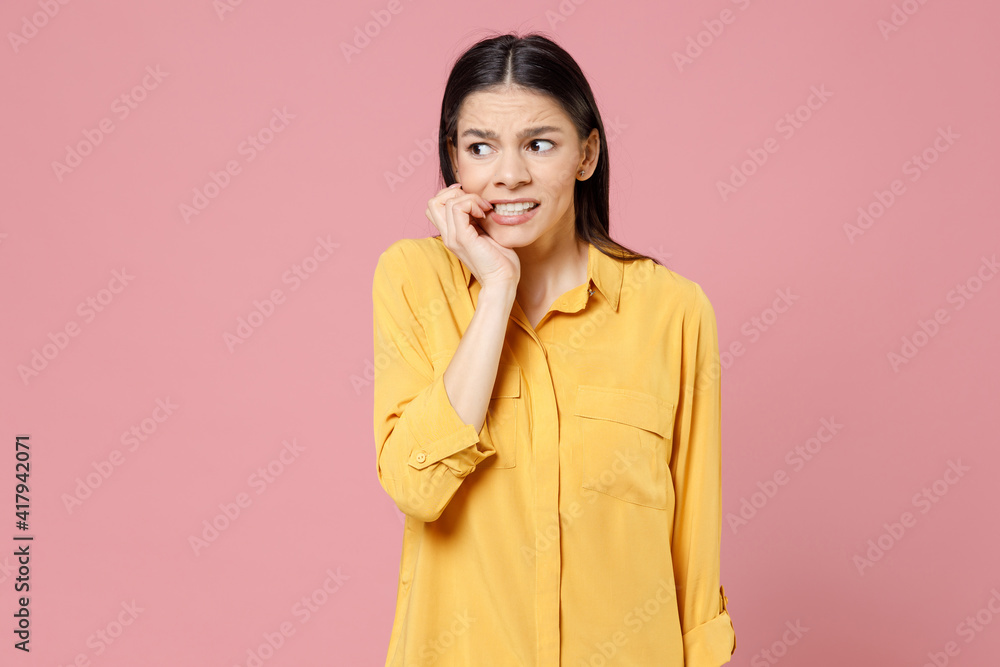 Young scared worried thoughtful pensive troubled brunette attractive latin woman 20s in yellow casual shirt biting lips nails fingers look aside isolated on pastel pink background studio portrait. Young scared worried thoughtful pensive troubled brunette attractive latin woman 20s in yellow casual shirt biting lips nails fingers look aside isolated on pastel pink background studio portrait.