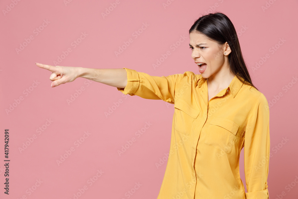 Young brunette cute serious strict latin student woman 20s wearing yellow casual shirt pointing index finger aside commanding scream shout isolated on pastel pink color background studio portrait. Young brunette cute serious strict latin student woman 20s wearing yellow casual shirt pointing index finger aside commanding scream shout isolated on pastel pink color background studio portrait.