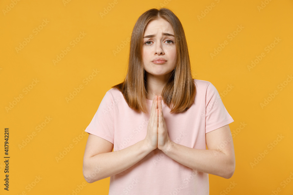 Young pleaded woman 20s with nude make up in casual basic pastel pink t-shirt, blank print design hands folded in prayer gesture, begging about something isolated on yellow background studio portrait Young pleaded woman 20s with nude make up in casual basic pastel pink t-shirt, blank print design hands folded in prayer gesture, begging about something isolated on yellow background studio portrait