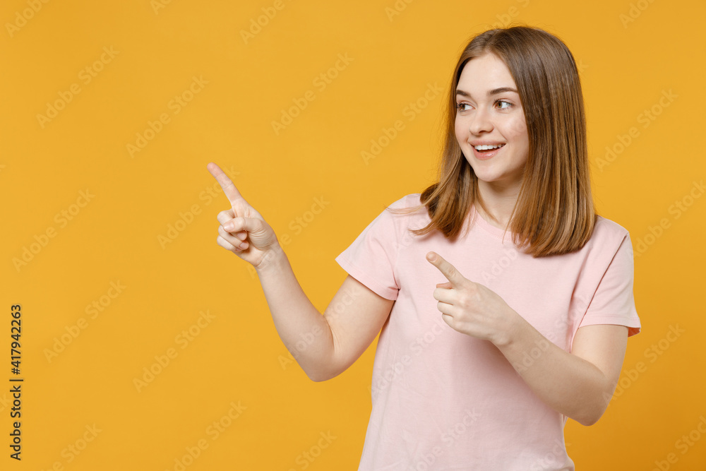 Young smiling happy positive woman 20s with nude make up wear casual basic pastel pink t-shirt pointing index finger forefinger aside on workspace area isolated on yellow background studio portrait Young smiling happy positive woman 20s with nude make up wear casual basic pastel pink t-shirt pointing index finger forefinger aside on workspace area isolated on yellow background studio portrait