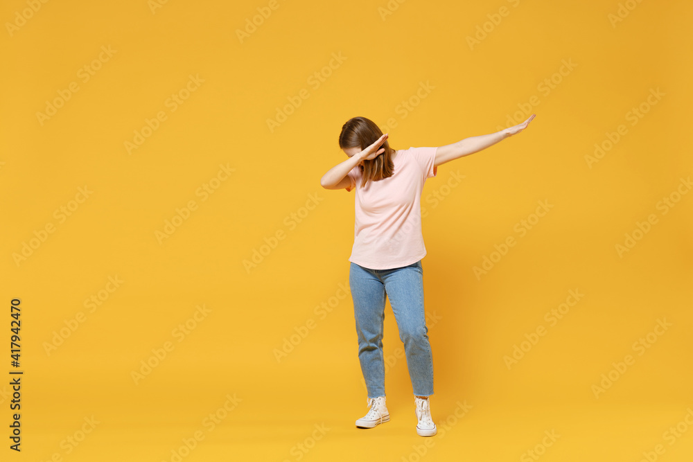 Full length of young woman in casual basic pastel pink t-shirt, jeans doing dab hip hop dance hands move gesture youth sign hiding covering face isolated on yellow color background studio portrait Full length of young woman in casual basic pastel pink t-shirt, jeans doing dab hip hop dance hands move gesture youth sign hiding covering face isolated on yellow color background studio portrait